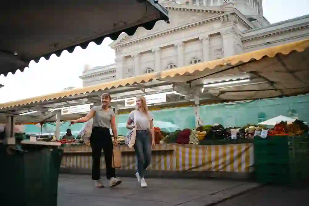 Entre les légumes frais et les étals colorés du marché, deux femmes se promènent dans la bonne humeur. L'atmosphère animée et la chaleur du matin confèrent à la scène une ambiance détendue et chaleureuse.