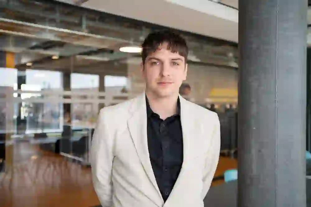 L'immagine mostra un uomo in un ufficio moderno di colore chiaro. Indossa una giacca bianca e una camicia nera, è in piedi e guarda direttamente nell'obiettivo. Lo sfondo mostra un ufficio aperto con pareti di vetro e un soffitto industriale. L'atmosfera è professionale e sicura e l'uomo dà un'impressione di calma e concentrazione.