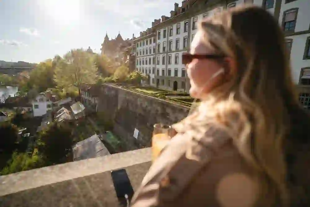 Woman enjoying a drink and city view from a bridge, with historic buildings and a castle in the background under a bright sky.