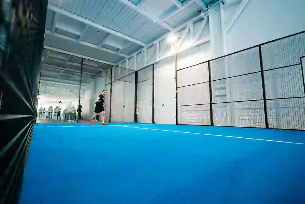 In the picture, a man is concentrating on playing padel on a blue indoor court. The scene radiates dynamism and sporting ambition. Spectators in the background add to the tension and focus of the game as the player prepares for the next shot.