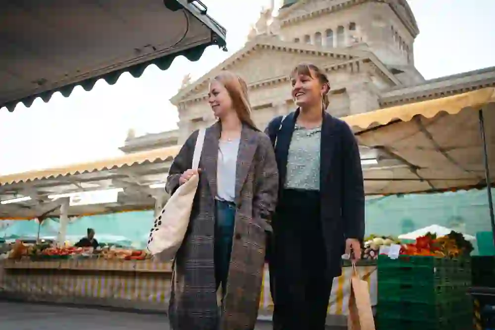 L'immagine mostra due donne che passeggiano davanti a una bancarella con le borse della spesa, apparentemente impegnate in una piacevole conversazione, mentre sullo sfondo si intravede un'architettura classica. L'atmosfera è rilassata e allegra.