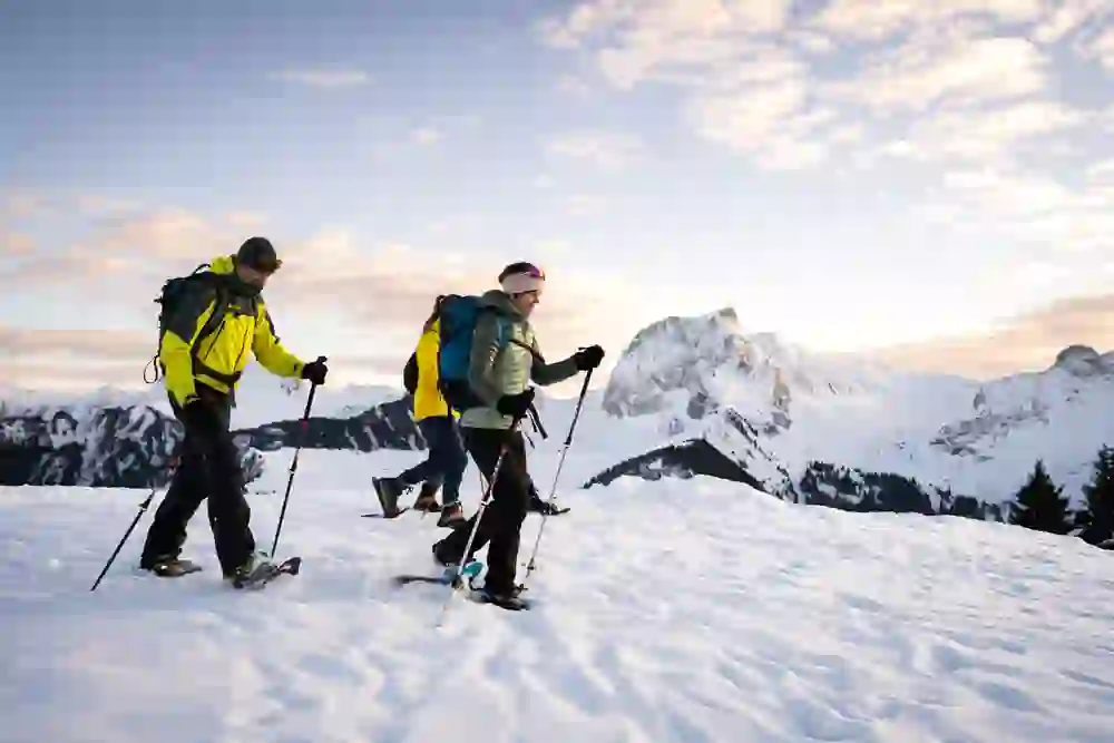 Das Bild zeigt eine Gruppe von Schneeschuhwanderern, die in der Dämmerung durch eine atemberaubende Winterlandschaft stapfen. Die schneebedeckten Berge im Hintergrund und der farbenfrohe Himmel erzeugen eine friedliche und zugleich aufregende Atmosphäre.