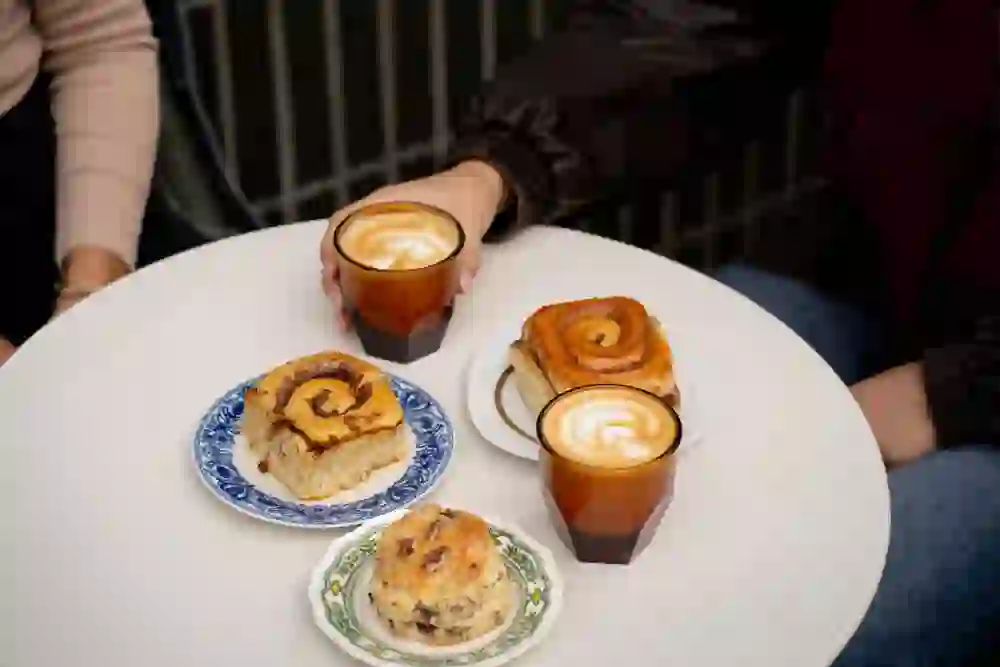 Auf dem Bild von Le Bread Sourdough sind frisch gebackene Zimtschnecken und duftende Cappuccinos auf einem runden Tisch zu sehen. Die warme Atmosphäre und das Zusammensein vermitteln Genuss, Gemütlichkeit und den perfekten Start in einen entspannten Tag.