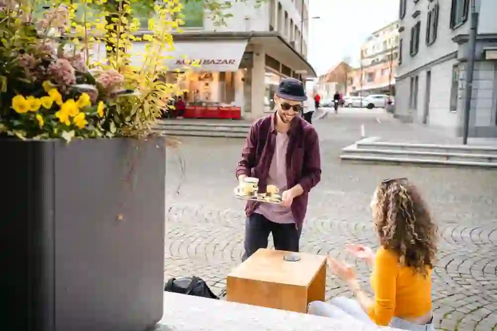 Ein Mann serviert mit einem Lächeln Kaffee an eine fröhliche Frau im Freien. Sie geniessen offensichtlich eine angenehme Unterhaltung in einer entspannten, urbanen Atmosphäre im Düby Spezialitäten Laden in Langenthal, umgeben von blühenden Pflanzen und der Lebhaftigkeit der Stadt.