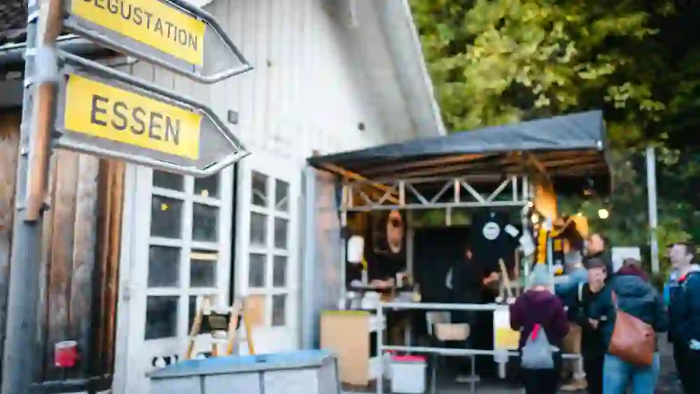 Das Bild zeigt zwei gelbe Schilder mit den Aufschriften "DEGUSTATION" und "ESSEN", die auf ein Gebäude hinweisen. Im Hintergrund ist ein Stand mit Menschen zu sehen, die sich unterhalten. Das Gebäude hat weiße Fenster und eine Holzwand, umgeben von grüner Vegetation.
