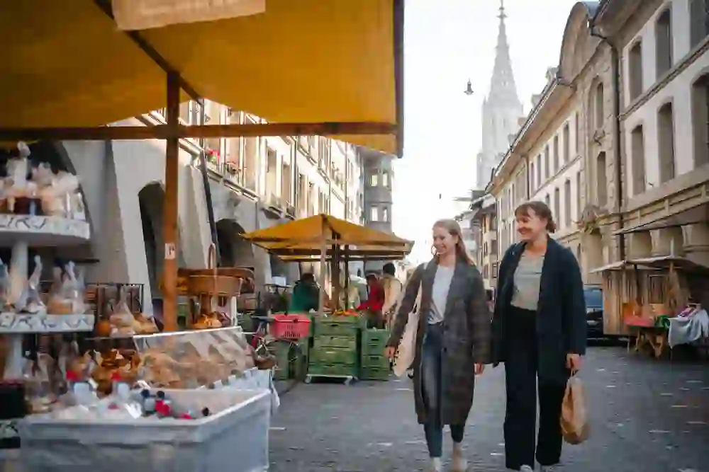 Auf dem Bild spazieren zwei Frauen lächelnd an Marktständen vorbei, die unter freiem Himmel in einer Strasse mit historischem Flair aufgebaut sind. Die Szene ist belebt und wirkt einladend.