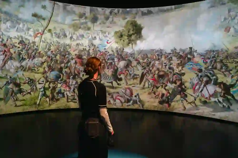 A visitor stands in front of a monumental circular projection of the Battle of Murten. The detailed panorama shows horsemen, flags and the turmoil of battle. The scene is overwhelming, dramatic and triggers feelings of wonder and contemplation.