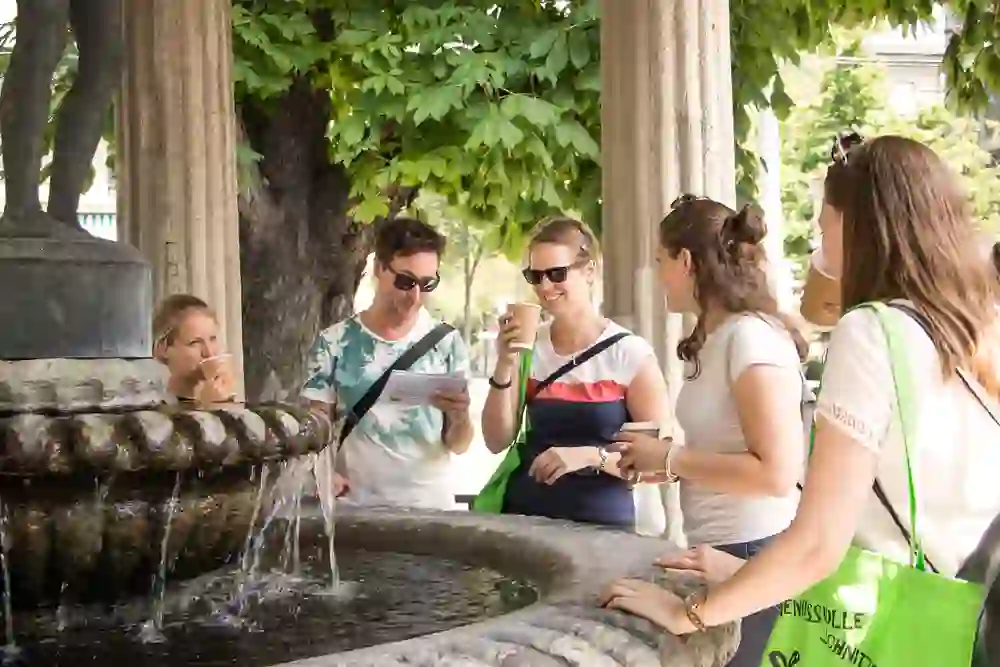Eine Gruppe lächelnder Menschen genießt bei Sonnenschein eine Pause an einem Brunnen, umgeben von grünen Blättern, mit Kaffee und eine Person liest etwas auf einem Dokument.