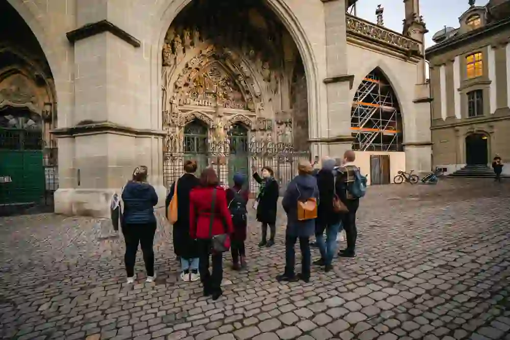 The photo shows a group of visitors standing in front of the entrance to the cathedral church, whose intricate stone latticework and filigree sculptures are impressive. The city guide in a black coat and green scarf stands in the foreground as she explains the history-laden building to the group. The cobblestones and historic architecture lend the picture an atmosphere of reverence and cultural interest.