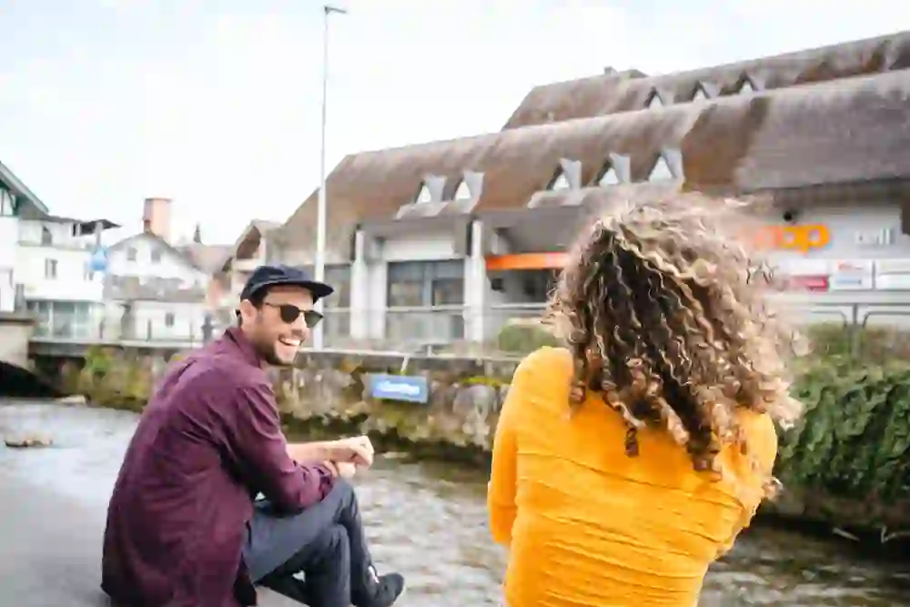 Ein Mann mit einem sonnigen Lächeln sitzt am Flussufer in der Innenstadt von Langenthal, während er sich mit einer Frau in leuchtendem Gelb unterhält. Ihr Rücken zum Betrachter, teilen sie eine scheinbar heitere und unbeschwerte Unterhaltung.