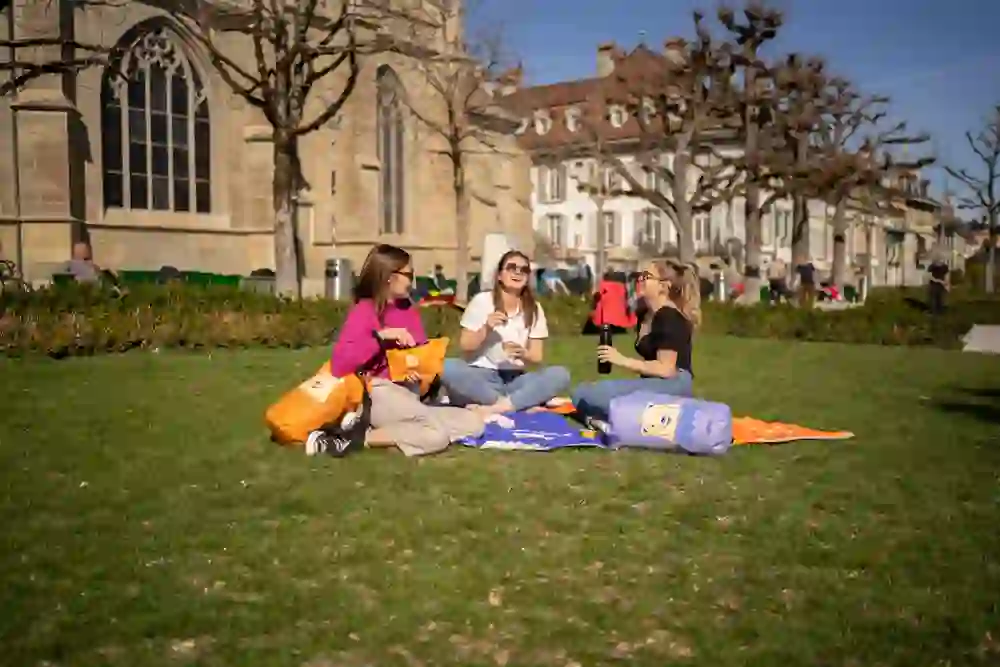 Auf der Münsterplattform in Bern geniessen drei Freundinnen ihre gemeinsame Zeit bei einem Picknick. Sie sitzen entspannt auf einer blauen Decke, umgeben von satten Grünflächen und der historischen Kulisse der Stadt. Das sonnige Wetter spiegelt die fröhliche Stimmung wider, während sie lachen, plaudern und sich erfrischen – ein Moment des einfachen Glücks und der Verbundenheit, eingefangen in einem friedvollen, städtischen Rückzugsort.