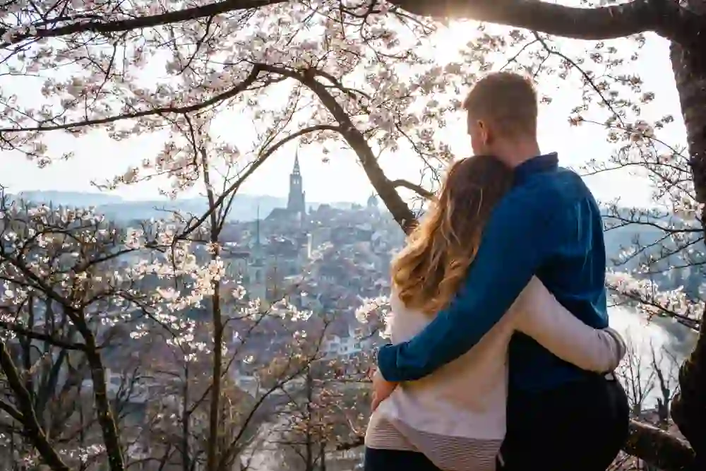 Un couple se tient dans la roseraie de Berne, entouré de la beauté délicate des fleurs de cerisier, et contemple la vieille ville historique avec sa cathédrale caractéristique. Le moment est rempli de romantisme et de silence, tandis qu'ils profitent ensemble de la vue pittoresque et des chauds rayons du soleil printanier.