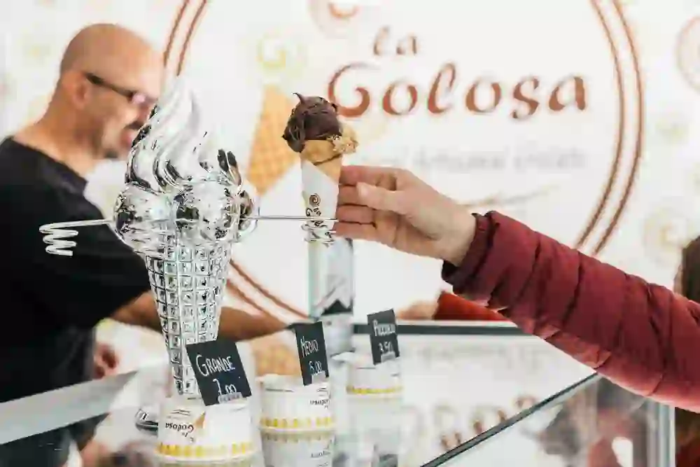 Ein Eis wird über die Theke der La Golosa Gelateria gereicht, mit einem verschwommen sichtbaren Verkäufer im Hintergrund. Die Szene strahlt Freude und Antizipation auf den Genuss des handwerklich hergestellten Gelatos aus.