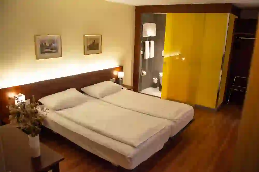 Das Bild zeigt ein gemütliches Hotelzimmer des Hotels Goldener Schlüssel mit zwei Einzelbetten, die zusammenstehen. Über den Betten hängen gerahmte Bilder. Die Beleuchtung ist warm und einladend, und die gelbe Glasschiebetür zum Badezimmer fügt einen lebhaften Farbtupfer hinzu. Auf einem kleinen Tisch steht eine Vase mit getrockneten Blumen, was zur wohnlichen Atmosphäre beiträgt.