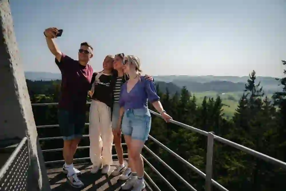 Vier Personen machen ein fröhliches Selfie auf einem Aussichtsturm mit Blick auf eine weitläufige, grüne Landschaft. Sie lachen und wirken entspannt. Die sommerliche Atmosphäre vermittelt Leichtigkeit und Freude am gemeinsamen Moment.