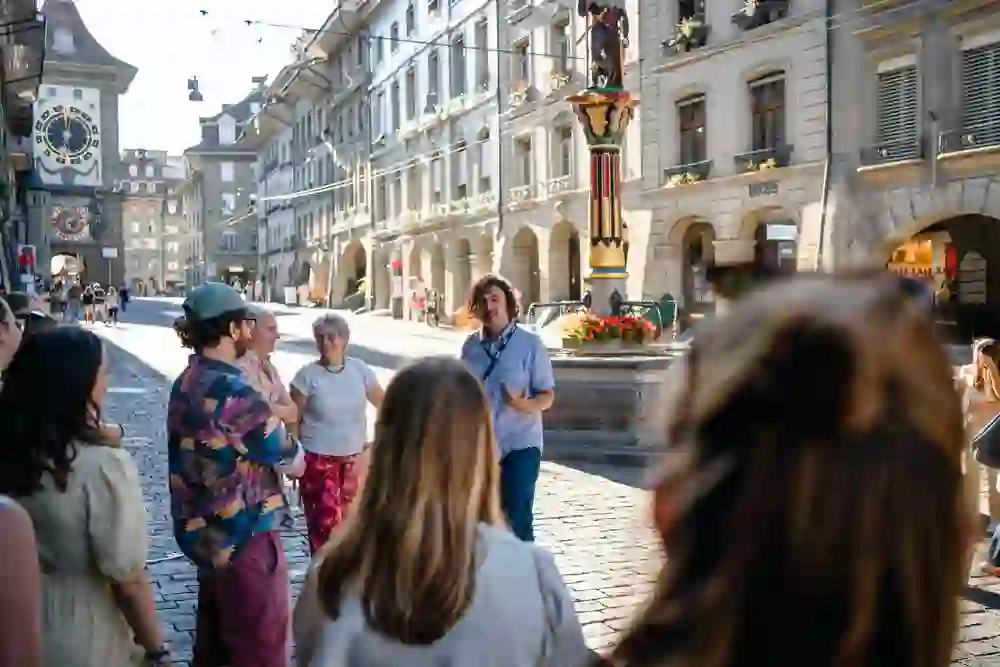 Das Bild zeigt eine Gruppe von Menschen, die einer Stadtführung folgen. Der Guide spricht gestikulierend zu den Teilnehmern. Im Hintergrund ist die Zytglogge (Zetglocke) und ein Brunnen in einer historischen Straße zu erkennen, wahrscheinlich in Bern.
