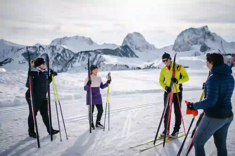 Vier Personen stehen in winterlicher Kleidung mit Langlaufskiern vor einer beeindruckenden Bergkulisse. Sie lächeln und scheinen sich auf den Skilanglauf vorzubereiten. Die Stimmung ist lebhaft, sportlich und von Vorfreude auf die Aktivität geprägt.