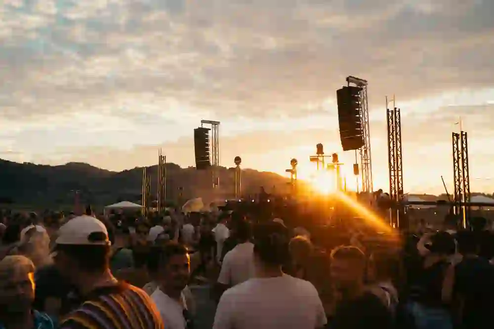 L'image montre une foule animée de festivaliers au coucher du soleil. Des personnes d'âges et de tenues différents profitent ensemble de la musique dans un environnement extérieur, avec en toile de fond de douces collines. L'éclairage chaleureux et le coucher de soleil confèrent à la scène une ambiance joyeuse et énergique.