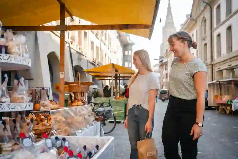 Zwei junge Frauen schlendern über den Markt in der Münstergasse in der historischen Berner Altstadt. Sie betrachten eine Auslage mit frischem Gebäck unter einem gelben Sonnendach. Im Hintergrund ist das Berner Münster zu sehen.