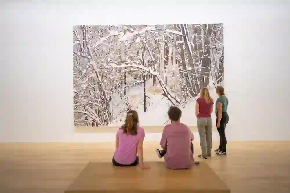 Vor einer riesigen, detailreichen Winterlandschaft im Museum Franz Gertsch verweilen vier Besucher*innen. Zwei sitzen auf einer Bank, zwei stehen staunend davor. Die Szene strahlt Ruhe und Kontemplation aus – ein Moment des Innehaltens in der faszinierenden Kunstwelt.