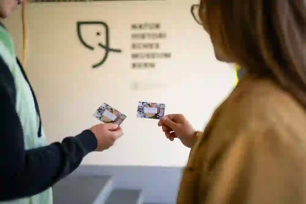 Zwei Personen stehen sich gegenüber und halten Museumskarten in die Kamera. Die unscharfe Museumswand im Hintergrund und die ruhige Geste des Austauschs vermitteln eine Atmosphäre der entspannten Erwartung.