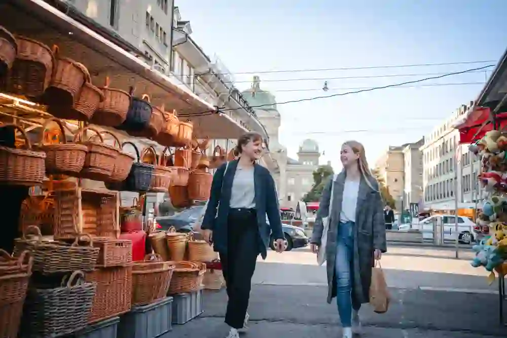 Das Foto zeigt zwei Frauen, die zwischen Marktständen am Waisenhausplatz entlanggehen. Links hängen zahlreiche geflochtene Körbe in verschiedenen Grössen und Farben. Rechts sind Plüschtiere an einem weiteren Stand zu sehen. Hinter den beiden Frauen ragt die Kuppel des Bundeshauses über den Gebäuden empor.