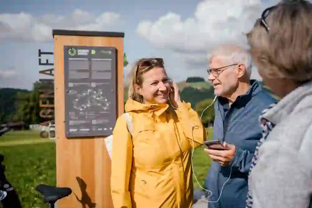 The picture shows two people listening to an audio tour of the "Gantrisch Legend Route" through headphones. The woman in the yellow mackintosh is laughing, while the man is smiling. The mood is cheerful and relaxed, full of anticipation for the adventure ahead.
