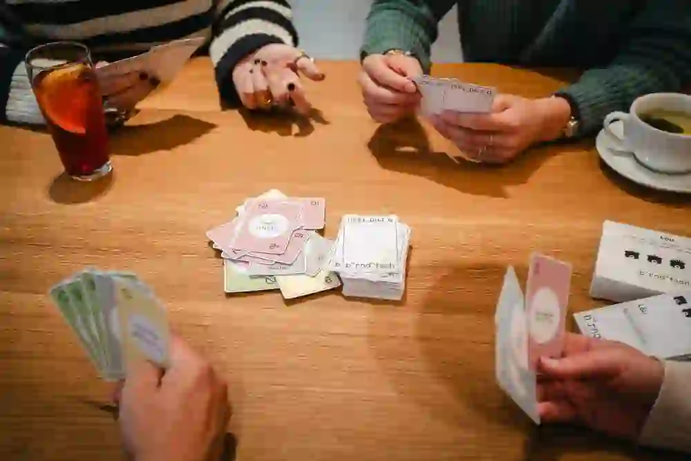 Das Bild zeigt vier Personen, die an einem Tisch ein Kartenspiel spielen. Die Karten sind in verschiedenen Farben und mit Berndeutsch-Wörtern beschriftet. Auf dem Tisch stehen ein Glas Eistee und eine Tasse Tee.