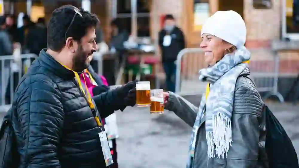 Das Bild zeigt zwei Personen, die mit Bierkrügen anstoßen. Der Mann links trägt eine schwarze Jacke und eine Sonnenbrille auf dem Kopf. Die Frau rechts trägt eine weiße Mütze, einen grauen Schal und eine graue Jacke. Beide lächeln. Im Hintergrund sind weitere Menschen und ein Zelt zu sehen. Die Szene scheint auf einem Außenfest stattzufinden.