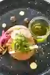 Auf dem Bild ist ein kunstvoll angerichtetes Gericht zu sehen: Ein fein zubereitetes Tatar, garniert mit frischen Radieschenscheiben, eingelegtem Gemüse, Kräutern und Pilzen. Dazu gibt es ein kleines Glas mit Olivenöl, dekorativ auf einem schwarzen Teller mit bunten Saucenpunkten und Knusper-Elementen serviert – modern, elegant und sehr appetitlich.