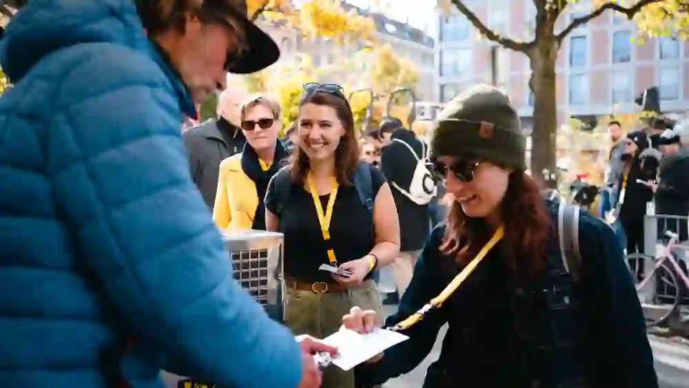 Auf dem Bild sind mehrere Personen bei einem Event im Freien zu sehen. Im Vordergrund reicht ein Mann in einer blauen Jacke etwas an eine Frau mit grüner Mütze und Sonnenbrille, die ein Lächeln trägt. Neben ihr steht eine weitere Frau mit schwarzem T-Shirt und Sonnenbrille auf dem Kopf, ebenfalls lächelnd. Im Hintergrund sind weitere Menschen zu sehen, darunter eine Person in einem gelben Mantel. Die Szene spielt sich in einer urbanen Umgebung mit herbstlich gefärbten Bäumen ab.