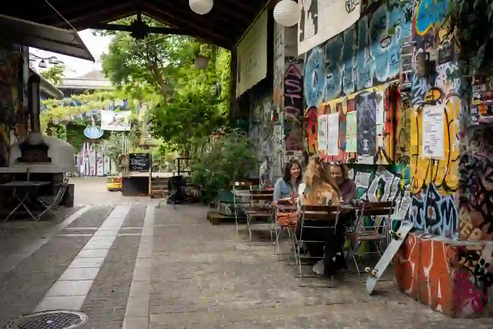 Three friends sit at a table in a charmingly overgrown courtyard, surrounded by colourful graffiti and urban art. The relaxed atmosphere invites you to linger and feel the vibrancy and creativity of the place.