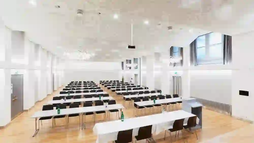 Der Saal im Kongresszentrum Kreuz Bern ist gross und hell, bereit für ein informatives Seminar. Die eleganten Leuchten und das strukturierte Deckendesign schaffen eine Atmosphäre der Erhabenheit und Intellektualität.