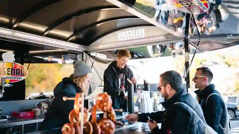 Das Bild zeigt einen Bierstand der Marke "Felsenau", an dem mehrere Personen bedient werden. Ein junger Mann zapft Bier, während eine Frau mit grauer Mütze im Hintergrund arbeitet. Zwei Männer im Vordergrund, einer mit Sonnenbrille, unterhalten sich und schauen auf ihr Handy. Im Vordergrund sind Brezeln auf einem Ständer zu sehen. Der Stand ist überdacht und spiegelt sich in einer glänzenden Oberfläche.
