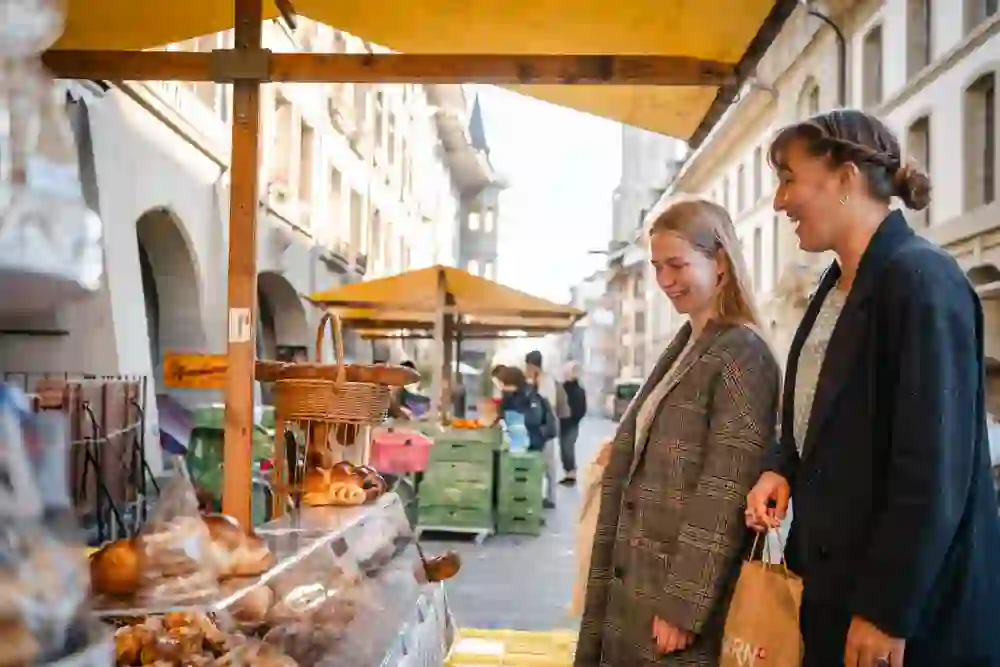 Due donne sorridenti guardano i prodotti da forno in una bancarella del mercato, immerse in una scena di trambusto urbano con un fascino storico sullo sfondo. L'atmosfera è allegra e spensierata.