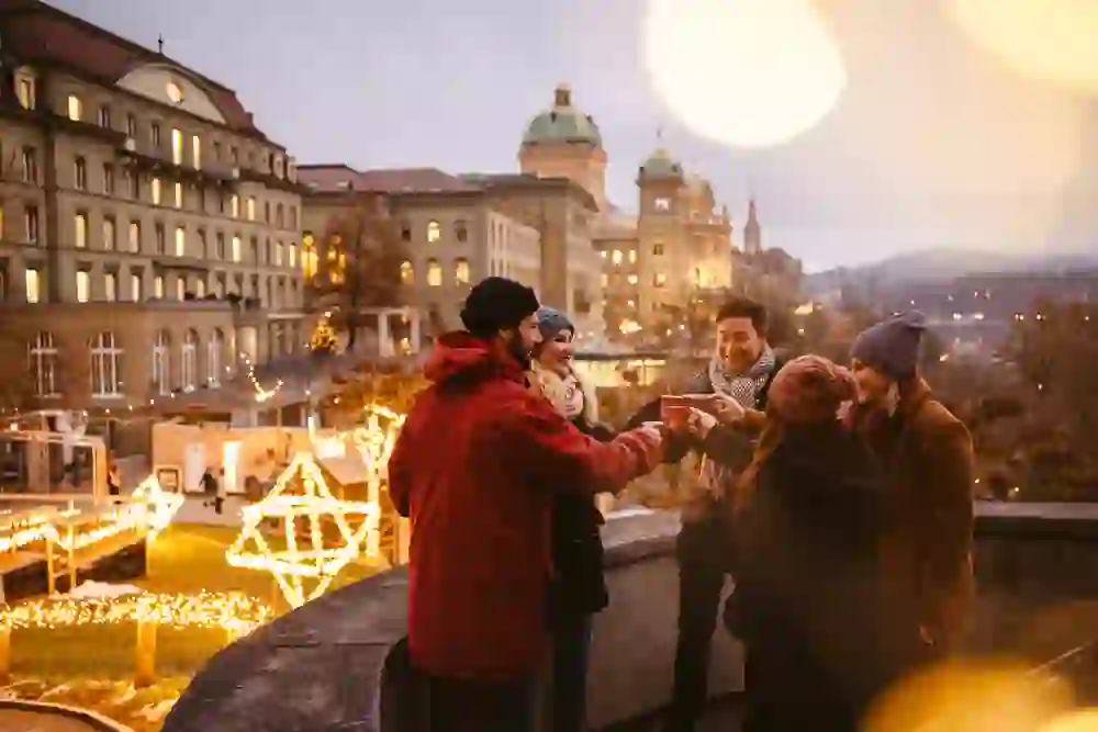 Das Bild zeigt eine Gruppe von Freunden, die auf einem weihnachtlich beleuchteten Platz in Bern zusammenkommen. Sie stoßen mit Tassen an und genießen die festliche Atmosphäre vor dem Bundeshaus. Die Stimmung ist warm und fröhlich, voller Gemeinschaftsgefühl und Vorfreude auf die Feiertage.