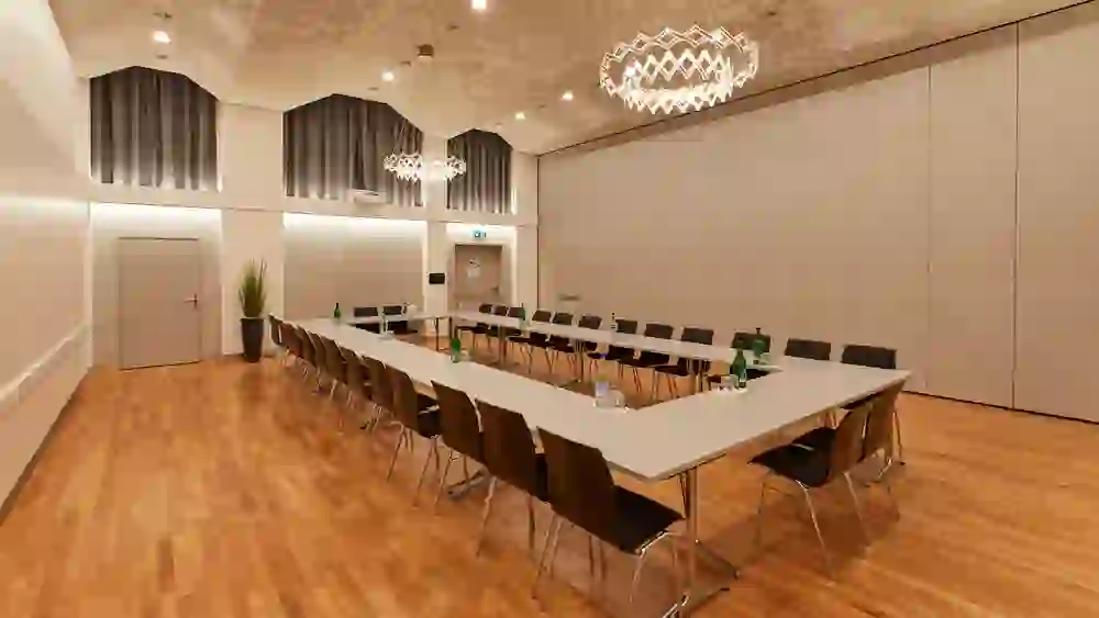 Der Saal im Kongresszentrum Kreuz Bern ist geräumig und stilvoll, mit einer langen Konferenztisch-Anordnung und edlen Leuchtern. Das moderne, doch behagliche Ambiente fördert eine produktive und angenehme Arbeitsatmosphäre.