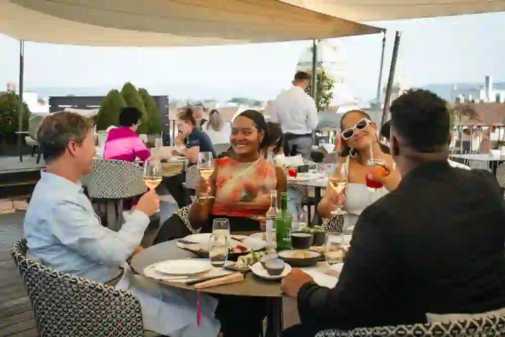 Sur l'élégante Sky Terrace, des amis profitent d'une soirée d'été détendue au-dessus des toits de Berne - avec de délicieuses boissons, un bon repas et une vue sur le Palais fédéral. L'ambiance est élégante et exubérante, marquée par le plaisir, la légèreté et la joie de vivre.