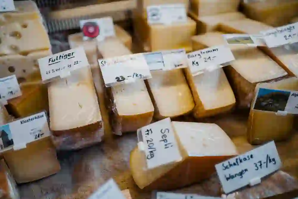 Das Bild präsentiert eine appetitliche Auswahl an Käsesorten von der Bergkäserei REK in Oberbütschel, sorgfältig beschriftet mit Namen und Preisen. Die verschiedenen Texturen und Farbtöne der Käsestücke, von cremigem Weiss bis zu reichhaltigem Gelb, zeugen von der Vielfalt und Qualität der Produkte. Die handgeschriebenen Etiketten verleihen der Präsentation eine persönliche und bodenständige Note, und man kann sich fast den reichen Geschmack und das Aroma vorstellen, das diese Käsesorten bieten.