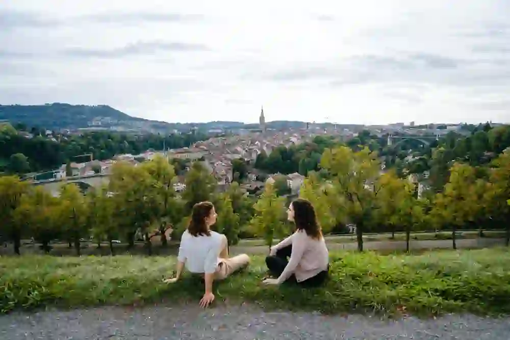 Two women sit relaxed in the grass of the rose garden in Bern, looking out over the city with its green trees and the striking cathedral in the background. It is a peaceful moment of friendship and reflection, with a view that captures the history and natural beauty of Bern.