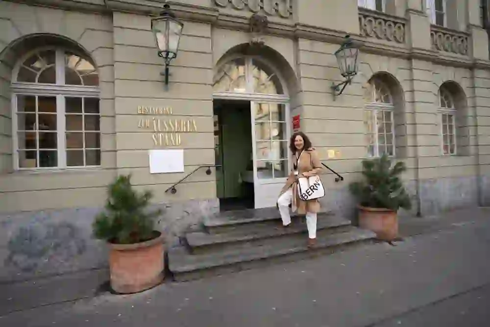 Eine fröhliche Frau posiert auf den Stufen eines eleganten Restaurants in Bern, ihr Lächeln fängt die Gelassenheit und den Charme der historischen Umgebung ein.