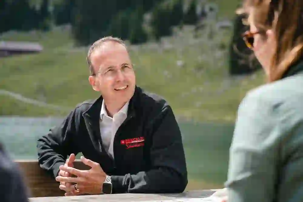 Auf dem Bild ist ein Gespräch in einer malerischen Bergkulisse zu sehen. Ein Mann im Gespräch lächelt entspannt, die Arme offen, während er draußen an einem rustikalen Holztisch sitzt, was eine warme, einladende Atmosphäre schafft.