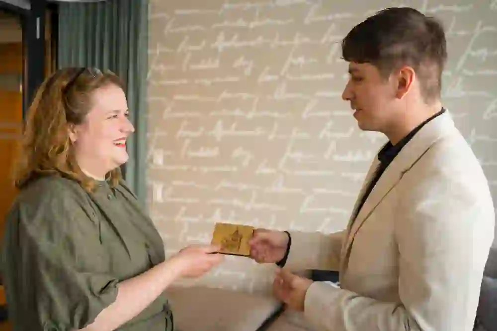 Auf dem Bild sind zwei Personen zu sehen, die sich gegenüberstehen und lächelnd eine hölzerne Tafel austauschen. Die Frau trägt ein grünes Kleid und lacht, während der Mann, der ein helles Sakko trägt, ebenfalls lächelt und die Tafel übergibt. Die Hintergrundwand ist mit handschriftlichen Schriftzügen dekoriert, was dem Raum eine persönliche und einladende Atmosphäre verleiht. Die Stimmung ist herzlich und freudig, was auf eine positive Interaktion zwischen den beiden Personen hinweist.