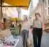 The picture shows two young women smiling as they stroll through a market in a historic alleyway. Surrounded by fresh baked goods and colourful market stalls, the scene conveys a friendly, relaxed and lively atmosphere.