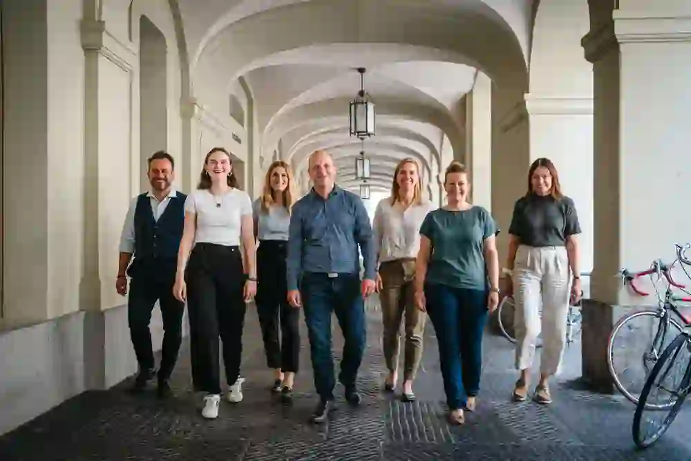 Un groupe de personnes souriantes marchent côte à côte sous les arches d'une tonnelle historique, exubérantes et au pas, ce qui donne une impression de cohésion et de dynamisme positif.