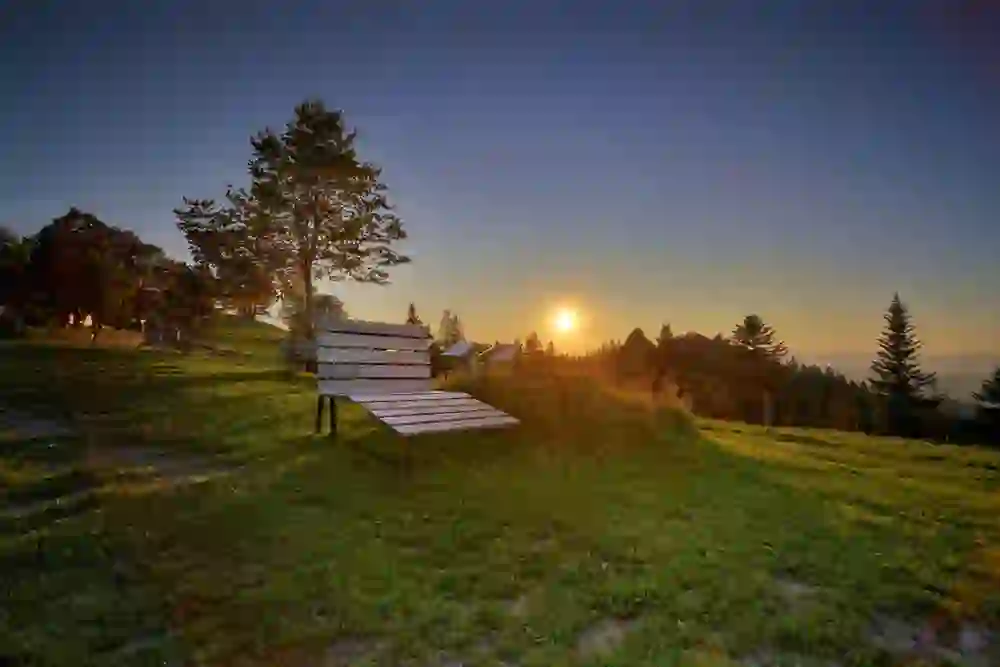 Das Bild fängt einen friedvollen Sonnenuntergang auf der Ahornalp im Emmental ein, mit dem warmen Glanz der Sonne, die gerade am Horizont niedergeht. Eine hölzerne Bank im Vordergrund lädt zum Verweilen ein, während die umliegenden Bäume und das weiche Abendlicht eine ruhige und erholsame Szene schaffen. Das Licht der untergehenden Sonne bringt die Landschaft zum Leuchten und schafft einen Moment der Stille und Schönheit.