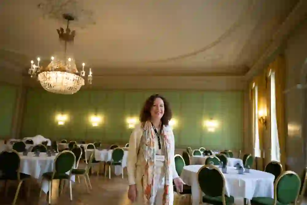 Une femme rayonnante se tient dans une salle somptueuse, entourée de lustres opulents et de tables élégamment dressées, qui dégagent une atmosphère d'histoire et de convivialité.