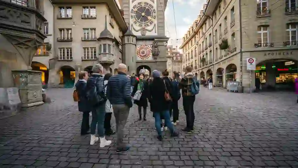 Sur cette photo, un groupe de personnes se tient sur une place pavée et observe la tour Zytglogge, qui est un élément caractéristique de la ville de Berne. L'ambiance est empreinte de culture et d'histoire ; les auditeurs sont plongés dans une explication donnée par la guide de la ville, qui se tient au centre. Il fait jour et la lumière éclaire doucement la scène, ce qui souligne le charme historique du lieu.