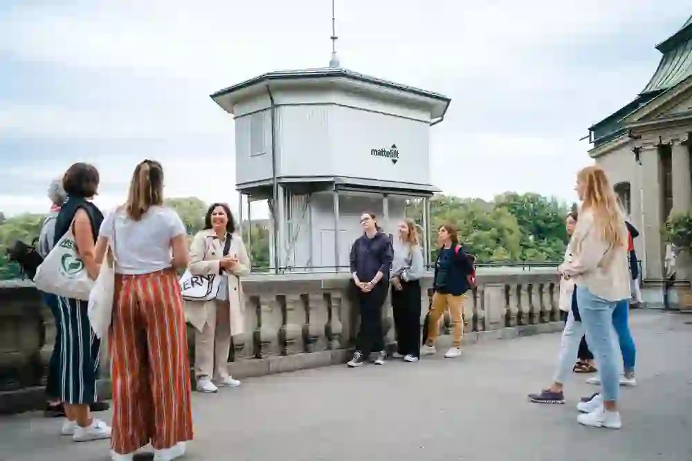 Eine Gruppe lauscht entspannt einer Führung ins historischen Berner Mattequartier, neben dem weissen Mattelift, umgeben von Grün.