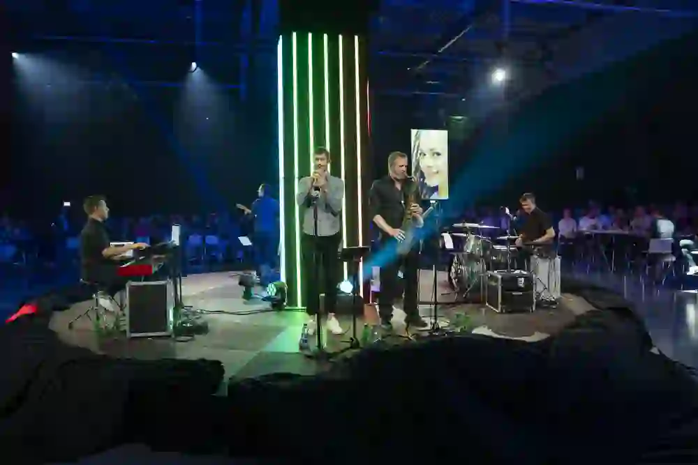 Eine Liveband spielt auf einer rund beleuchteten Bühne, umgeben von sitzendem Publikum. Leuchtende Farben, intensive Klänge und gespannte Gesichter erzeugen eine mitreissende, lebendige Atmosphäre voller Energie, Nähe und gemeinsamer Begeisterung.